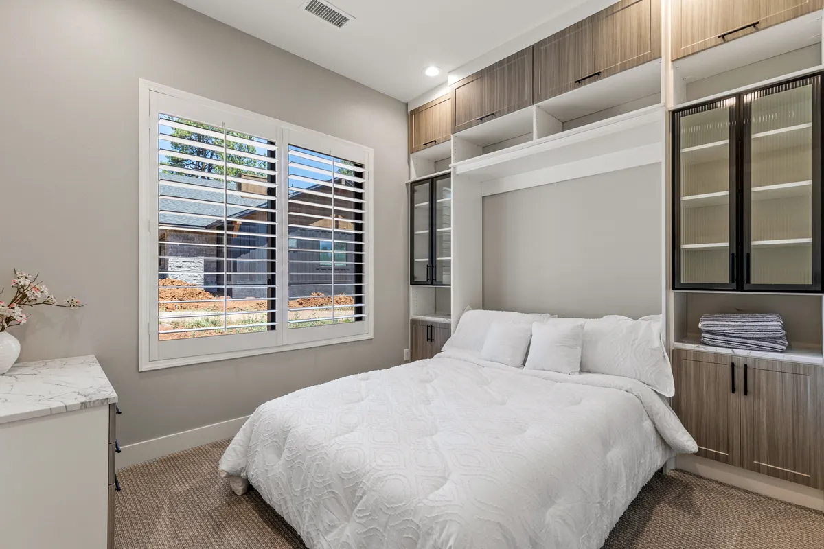A tranquil bedroom featuring a bed with crisp white bedding, a large window with shutters allowing plenty of natural light, and built-in cabinetry. The space is inviting and restful, providing a sense of privacy and comfort.