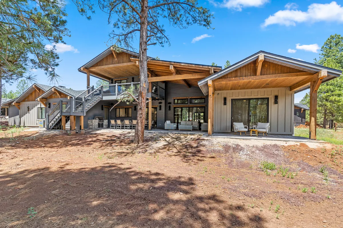 A large wooden deck extends from the house, surrounded by towering pine trees and featuring expansive windows that blur the line between indoor and outdoor living. The deck is furnished with seating areas for dining and relaxation, offering views that invite guests to unwind or gather at sunset.