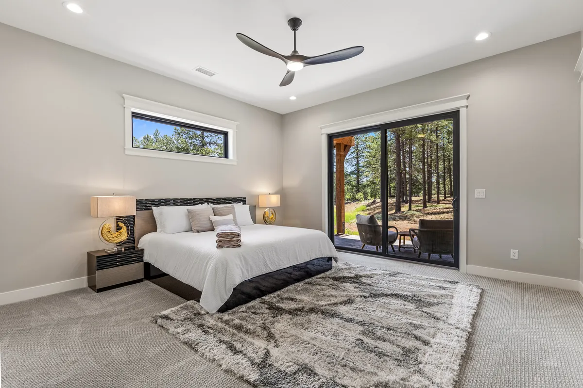 A serene bedroom featuring a comfortable bed with plush white linens, a sliding glass door opening to a patio, elegant nightstands, a ceiling fan, and a cozy area rug. Soft natural light fills the room, emphasizing its tranquil, resort-like atmosphere.