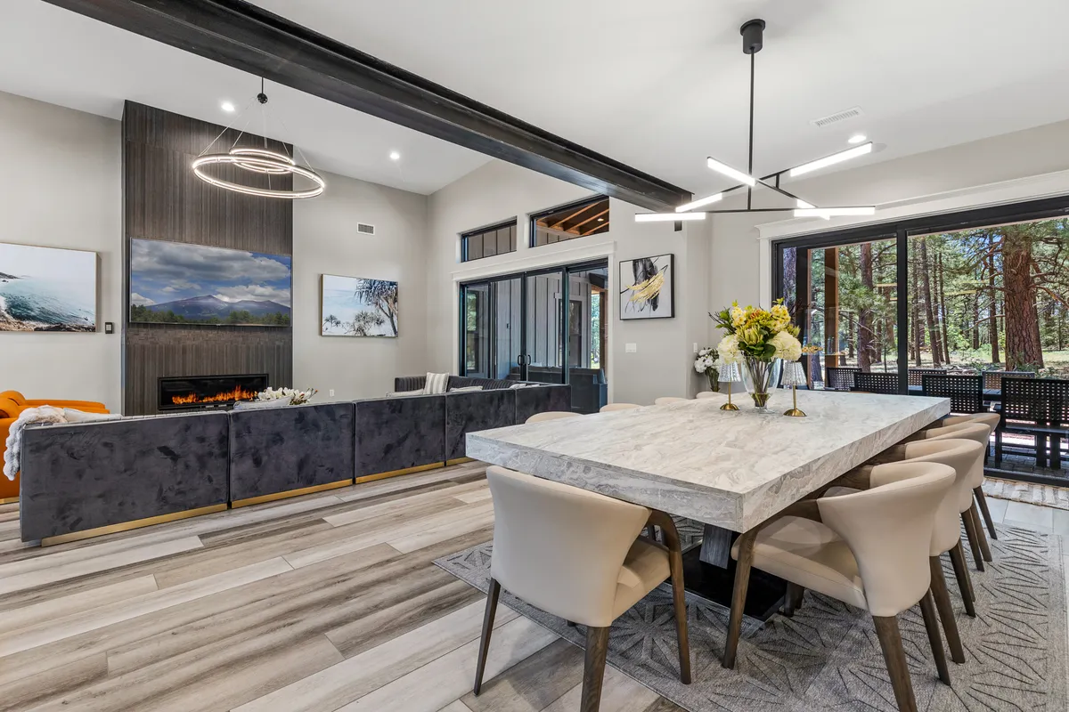 This image captures a spacious open-concept living and dining area with a large dining table, comfortable sofa, artwork on the walls, and expansive windows that flood the space with natural light. The atmosphere is warm and inviting, with the fireplace offering a cozy focal point. The space is clearly designed for group gatherings, family meals, and enjoying the surrounding views.