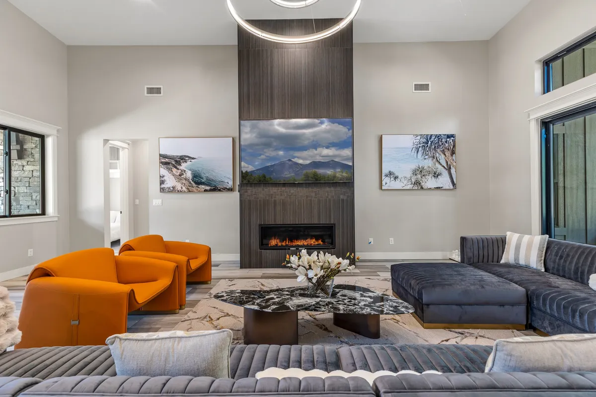The image showcases a spacious, elegantly decorated living area with a large, inviting sectional sofa gathered around a sleek modern fireplace. Stylish lighting, contemporary artwork, and expansive windows create a warm and sophisticated atmosphere, perfect for socializing or unwinding after a day in the Flagstaff pines.