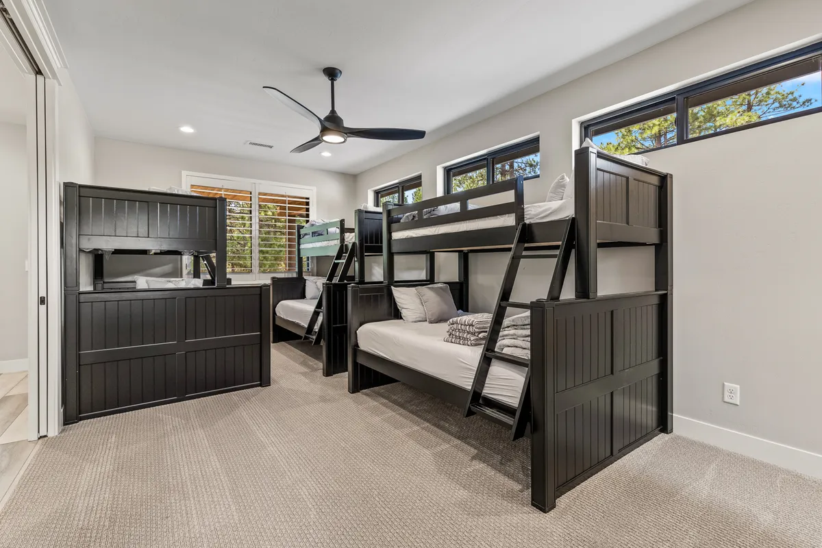 A playful and spacious bunk room with three twin-over-queen bunk beds, a ceiling fan, and bright windows. The setting is perfect for kids or groups of friends, offering space for both sleeping and social activities like reading or playing games.