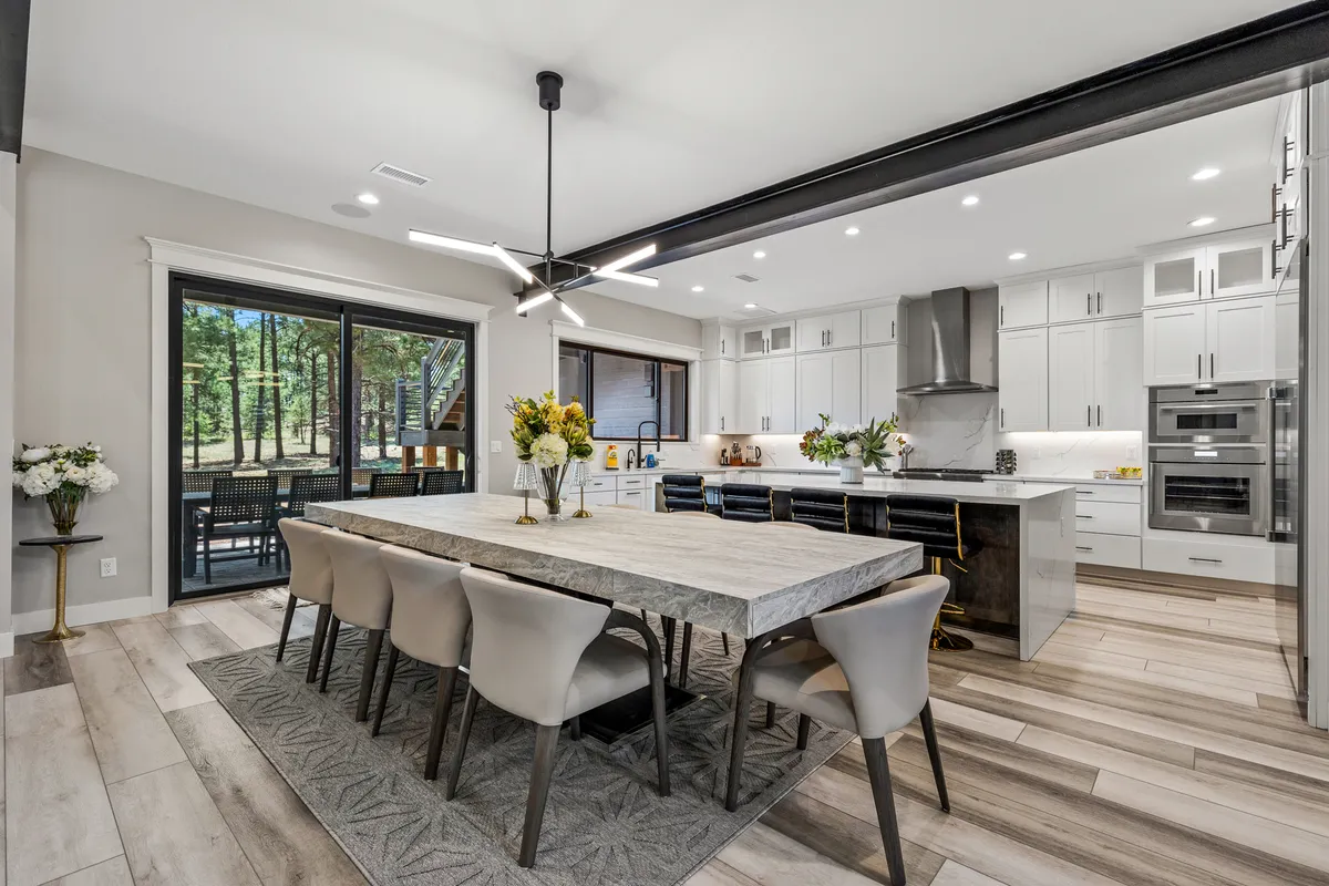 A bright, communal space featuring a large dining table adjacent to a modern kitchen. The area is filled with natural light from surrounding windows, and modern kitchen appliances—including what could be an ice machine and wine cooler—are visible, creating a perfect setup for entertaining and shared meals.