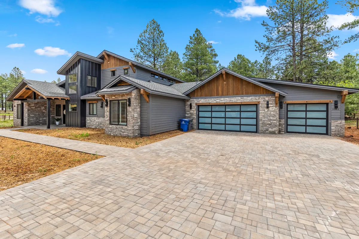 A wide view of the home's modern architectural design, featuring a stone and wood facade, a spacious driveway, and a serene yard surrounded by tall trees. The setting conveys an inviting, nature-immersed atmosphere, ideal for those looking to explore and adventure outdoors. Multiple parking spaces are visible, hinting at the convenience for guests or adventurers with extra vehicles or equipment.